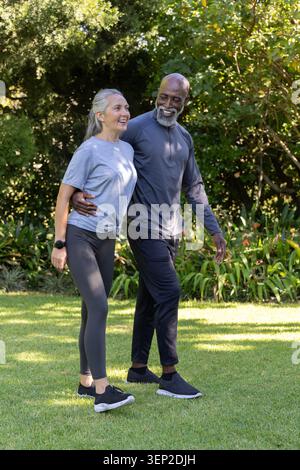 Senior couple walking arm-in-arm across sunlit park lawn, wearing athletic shoes and wristwatch. Garden, outdoors, greenery, flowers, lifestyle, compa Stock Photo