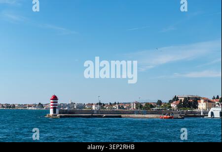 A distinctive red and white striped lighthouse beacon stands on a concrete pier against a bright coastal town backdrop. Stock Photo
