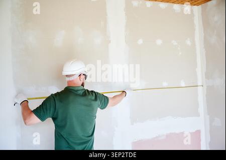 Builder measuring wall width with tape for layout planning. Worker in helmet marking alignment for installation, concept of precision work, interior renovation and construction preparation Stock Photo