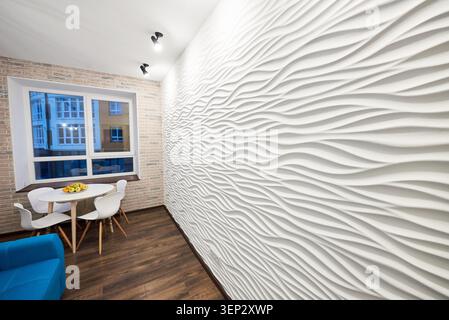 Dining area beside large 3D wave wall in modern apartment interior, with round table and chairs by window. Stylish decorative finish, brick accent and cozy urban living concept Stock Photo