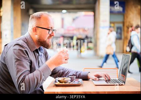 Man drinking juice while using laptop at outdoor cafe, smiling during work break, combining refreshment and remote tasks, concept of flexible work lifestyle in urban setting Stock Photo