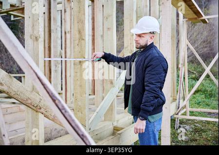 Builder using measuring tape to check gap between timber posts inside unfinished house structure, ensuring correct positioning and proportions on construction site Stock Photo