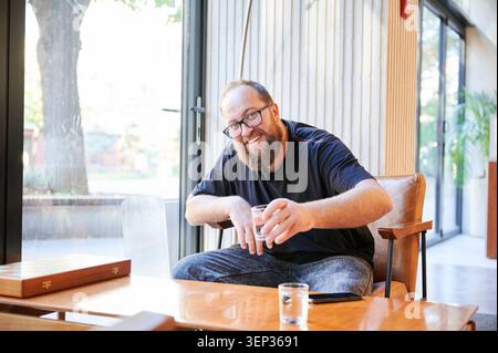 Smiling man sits in modern living room and holds glass of water near coffee table. Cozy indoor lifestyle scene for home comfort, relaxation, hospitality, or real estate promotion Stock Photo