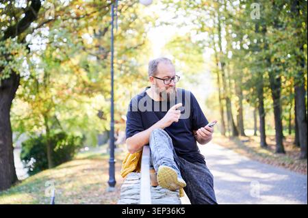 Relaxed bearded man in park. Sunlit park with man using his phone. Casual man with beard checks phone in outdoor setting. Laidback man with beard holds phone on park bench among autumn leaves Stock Photo