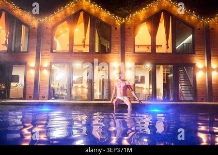 Man sitting on edge of outdoor pool at night in front of modern illuminated wooden cabins, enjoying warm water and cozy ambiance, luxury wellness and holiday retreat concept Stock Photo