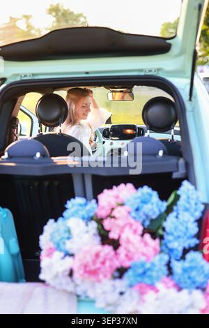 Smiling young woman sitting inside a pastel car with colorful hydrangeas in the trunk during golden hour, enjoying a countryside road trip Stock Photo