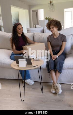 Two women of African descent sitting on sofa at home holding shipping box and tablet Stock Photo