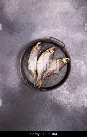 Three dried fish, known as vobla or taranka, are lying on a rustic metal tray, presenting a traditional seafood snack from an overhead perspective on Stock Photo
