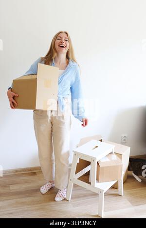 Happy young woman carrying a cardboard box into a new home, celebrating relocation, fresh beginnings and modern independent living. Stock Photo