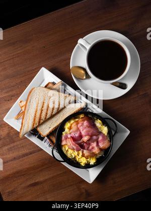 Warm breakfast scene with coffee and toast. Scrambled eggs with ham sit in black skillet. Golden-brown toast rests on white plate. Coffee cup, spoon, Stock Photo
