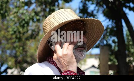 Older ukrainian man wearing in vyshyvanka talking on mobile phone at summer garden. Senior male in straw hat using cell phone outdoor. Concept of Stock Photo