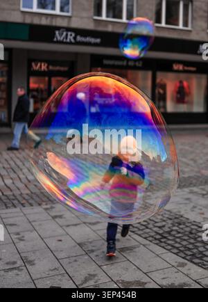 Child reflected in giant iridescent soap bubble floating on city street on an autumn day, Tallinn, Estonia. Stock Photo