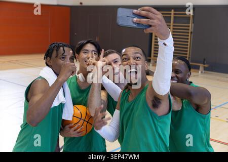 Diverse male teammates wearing green jerseys taking selfie with phone on court holding basketball Stock Photo