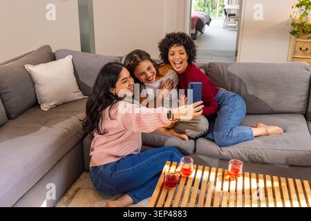 Diverse female friends taking selfie on gray sectional couch at home with smartphone Stock Photo