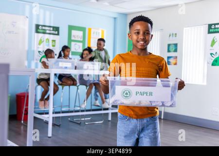African American boy wearing orange tee and jeans holding clear bin labeled PAPER in classroom Stock Photo