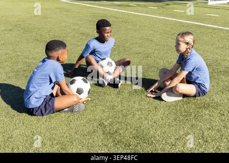 Three children sitting and talking on soccer pitch wearing matching blue shirts with two balls Stock Photo