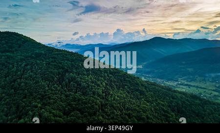 Evening drone scene of forested mountains fading into a blue valley. Warm light touches ridges above a scattering of villages while layered clouds col Stock Photo