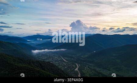 Sunset light glows over a winding river that threads through a broad mountain valley. Mist curls along the slopes as towering clouds rise above villag Stock Photo