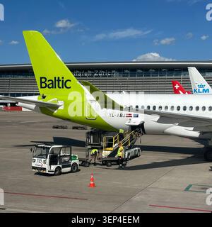 Swissport ground crew unload baggage from an airBaltic Airbus A220-300 during turnaround operations at a passenger gate with terminal in background. Stock Photo