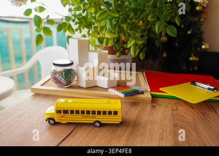 Yellow school bus toy on wooden desk with markers paper clips notebooks and green plant for back to school education concept homeschool workspace Stock Photo