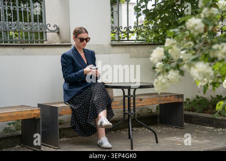 Young woman in dark blazer and sunglasses sits at outdoor terrace surrounded by greenery and flowering branches, looking upward in quiet courtyard Stock Photo