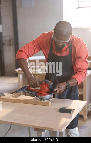 Woodworker smoothing wooden plank with electric hand sander at workshop bench, with window light Stock Photo