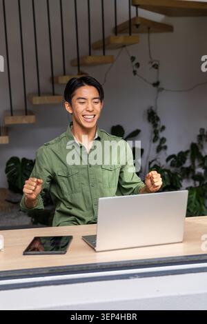 Asian man sitting at home desk using laptop, raising fists and smiling, wearing green shirt Stock Photo