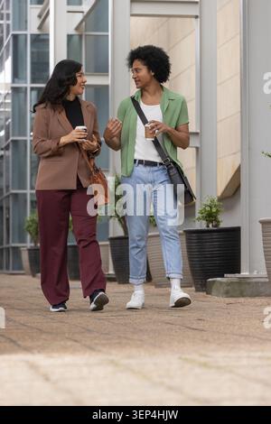 Diverse business-casual coworkers walking and talking on office path, holding coffee cups and bags Stock Photo