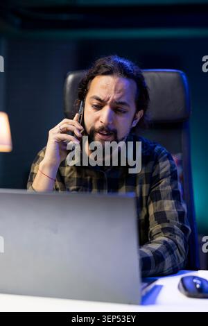 Multitasking individual efficiently manages a conversation on his mobile device while simultaneously working on digital laptop. Male person having remote communication with clients on cell phone. Stock Photo