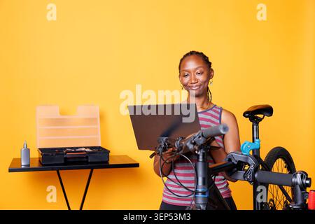 In front of yellow background, black woman performs bike maintenance by utilizing digital laptop for instructions. African american female using minicomputer and professional tools for fixing bicycle. Stock Photo