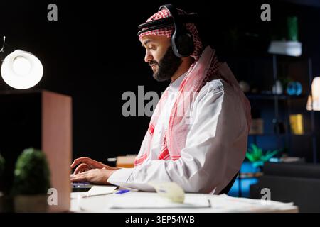 Side-view shot of a handsome Arab businessman works in modern office, using wireless technology and traditional attire. He engages in video calls while browsing, researching, and communicating online. Stock Photo
