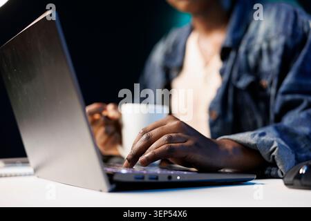 Detailed shot of african american individual holding a cup while working on the laptop. Black person with coffee mug typing on wireless computer, researching, studying online. Stock Photo