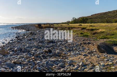 A coastal landscape features a rugged shoreline covered in smooth, light-colored stones on the island of Mykonos, Greece Stock Photo