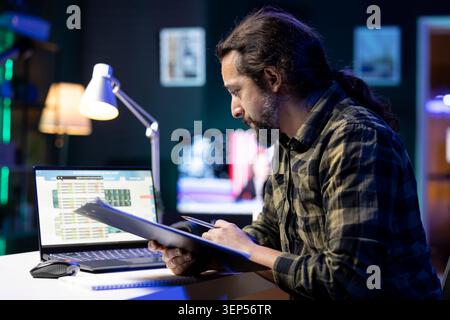 Young man works from home, using his laptop and taking notes on a notepad at a modern desk in his apartment. Male individual writing on a notebook, studying and researching financial data. Stock Photo