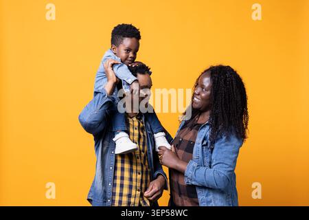 Sweet happy family with a small toddler posing in front of the camera, father carrying his son on his shoulders in studio while African American parents feeling grateful for their baby boy. Stock Photo