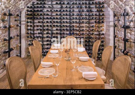 A long wooden dining table set for a formal dinner inside a rustic stone wine cellar with extensive bottle racks in Saint-Paul-de-Vence, France Stock Photo