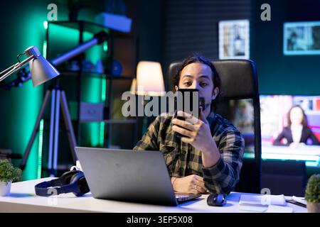 Portrait of a young man using his mobile device to communicate with friends while seated in front of laptop while young guy is holding his smartphone and using social media and the internet. Stock Photo