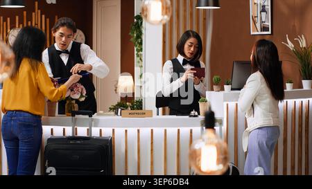 Tourists going through check-in process at hotel reception desk, staff verifying passport id for registration, two women signing ledger and reservation papers for accommodation on trip. Stock Photo