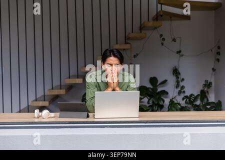 Asian man wearing olive shirt sitting behind counter at home using laptop tablet with headset Stock Photo