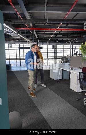 Two middle-aged men in blue shirt and mustard jacket walking, talking, holding tablet in office Stock Photo
