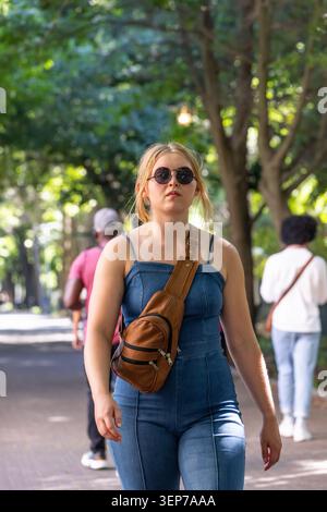 Woman walking on tree-lined path, wearing denim jumpsuit, round sunglasses, carrying sling bag Stock Photo