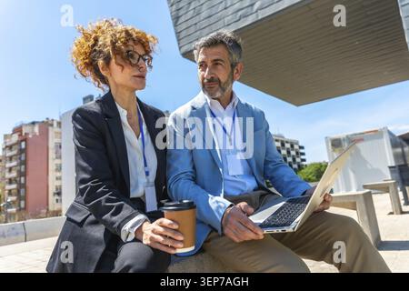 Two diverse business professionals sitting outdoors, discussing work and collaborating while using a laptop and drinking coffee with identification ba Stock Photo