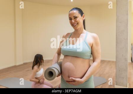 Pregnant woman smiling at camera in a bright studio, holding a rolled yoga mat and touching her belly while preparing for prenatal exercise and mindfu Stock Photo