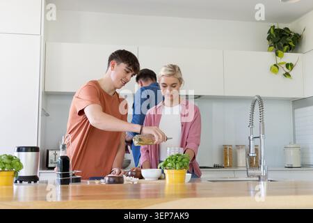 Housemates preparing dinner in modern kitchen, youth pouring olive oil from glass bottle Stock Photo