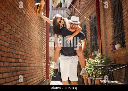Happy interracial couple having fun piggyback ride in Tbilisi old city narrow street.Cheerful young man giving girlfriend piggyback ride.Couple laughi Stock Photo