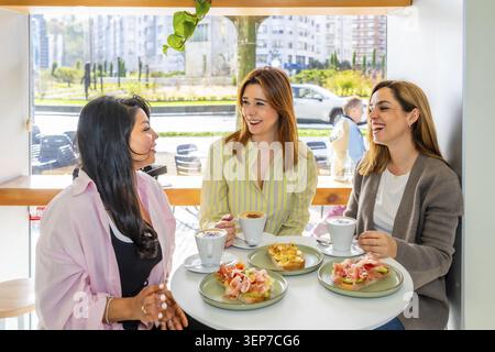 Three smiling women are having an engaging conversation while enjoying coffee and gourmet toasts, sharing a pleasant breakfast time together in a brig Stock Photo