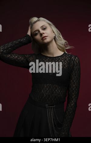 A stylish woman stands confidently, wearing a black lace top. Her long hair falls softly around her shoulders as she poses against a rich red backdrop Stock Photo