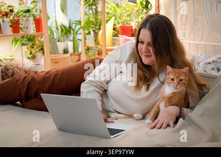Woman works remotely from her cozy bed with a laptop, while an affectionate red cat sits comfortably on her lap. The warm atmosphere includes many pla Stock Photo