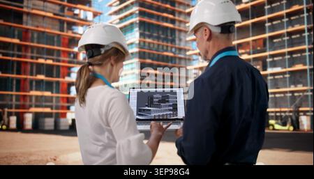 Two Architects And Engineers Discuss 3D BIM Model At Busy Construction Site. Stock Photo
