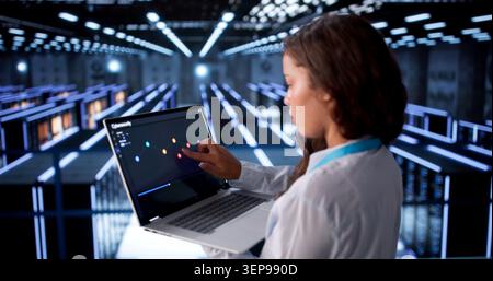 Woman Engineer Monitors Data Servers Using Laptop In Busy Cloud Center. Stock Photo